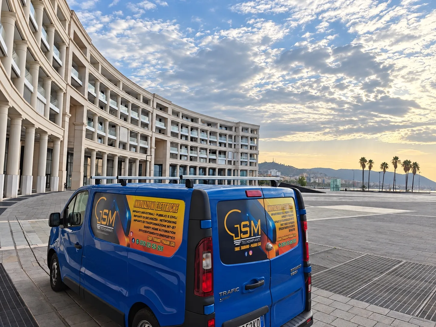 Il furgone blu di G.S.M. Tecnoimpianti con l'adesivo dell'azienda, in una piazza con un palazzo a semicerchio e le palme